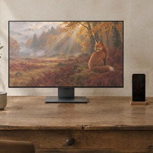 May include: A computer monitor displaying a digital painting of a red fox sitting in an autumn forest with golden sunlight filtering through the trees. The monitor is placed on a wooden desk next to a ceramic mug and a smartphone on a stand.