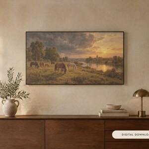 May include: A rectangular landscape painting in a thin black frame depicting a group of horses grazing in a grassy field near a river at sunset. The scene features a cloudy sky, distant trees, and a building silhouette under a warm golden light.