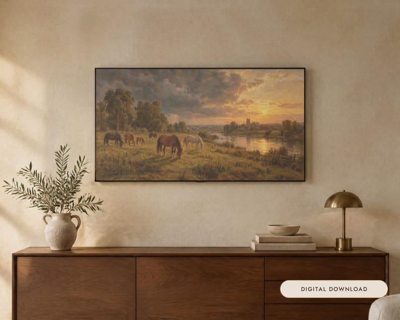 May include: A rectangular landscape painting in a thin black frame depicting a group of horses grazing in a grassy field near a river at sunset. The scene features a cloudy sky, distant trees, and a building silhouette under a warm golden light.