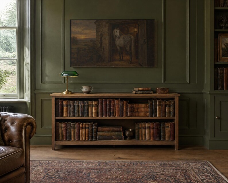 May include: A wooden bookcase with two shelves containing a collection of antique leather-bound books. Positioned on top is a green glass banker's lamp with a brass base and a small ceramic bowl. Above the unit is a framed painting of a horse.
