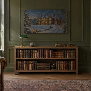 K&ouml;nnte beinhalten: Ein h&ouml;lzernes B&uuml;cherregal mit zwei Ebenen voller alter B&uuml;cher, gekr&ouml;nt von einer gr&uuml;nen Bankerlampe und einer kleinen Schale. &Uuml;ber dem Regal h&auml;ngt ein gerahmtes Landschaftsgem&auml;lde eines gro&szlig;en Herrenhauses im n&auml;chtlichen Schnee.