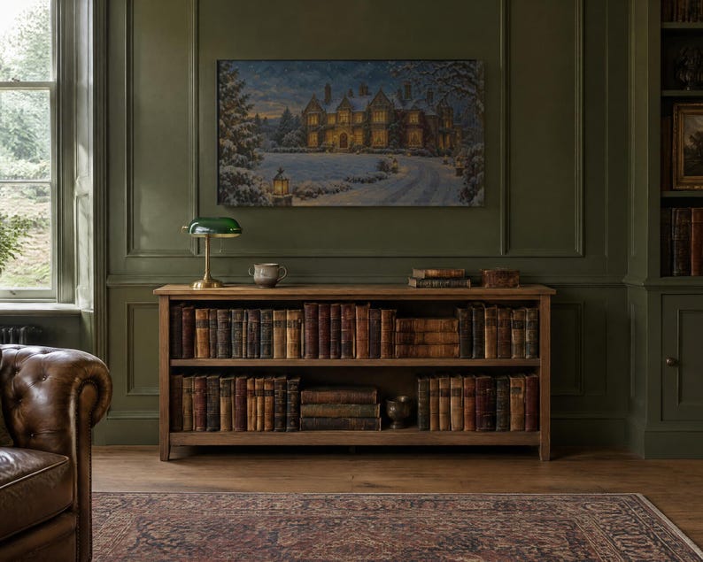 K&ouml;nnte beinhalten: Ein h&ouml;lzernes B&uuml;cherregal mit zwei Ebenen voller alter B&uuml;cher, gekr&ouml;nt von einer gr&uuml;nen Bankerlampe und einer kleinen Schale. &Uuml;ber dem Regal h&auml;ngt ein gerahmtes Landschaftsgem&auml;lde eines gro&szlig;en Herrenhauses im n&auml;chtlichen Schnee.