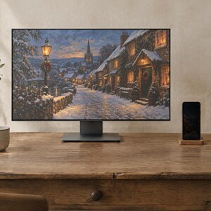May include: A computer monitor displaying a winter village scene with snow-covered stone cottages, a lit street lamp, and a church steeple at dusk. The monitor sits on a wooden desk next to a smartphone on a stand and a ceramic mug.