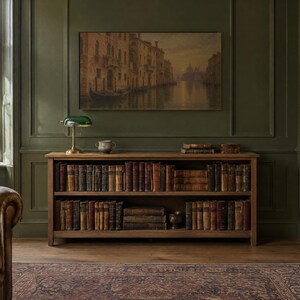May include: A wooden bookshelf with two tiers filled with antique leather-bound books. The unit features a rectangular frame and sits against a dark green paneled wall beneath a large landscape painting of a canal scene and a green banker lamp.