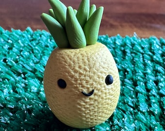 May include: A small, yellow pineapple with a green leafy top and a smiling face. The pineapple is sitting on a green, artificial grass surface.