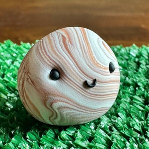 Puede incluir: Una figura de arcilla redonda y caprichosa con una cara sonriente. La figura tiene un patrón veteado en tonos de blanco, marrón y naranja. Puntos negros forman los ojos y una línea negra curva crea la sonrisa. Descansa sobre césped artificial verde.