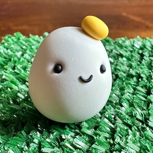 Puede incluir: Una figurita blanca en forma de huevo con una cara sonriente y ojos negros. La figurita tiene un pequeño círculo amarillo en la parte superior de su cabeza.