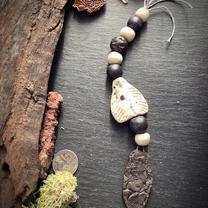 May include: A handcrafted necklace with ceramic beads in black, cream, and brown. The necklace includes a textured, dark grey pendant and a cream-coloured, wing-shaped bead. It is strung on a thin, off-white cord. The necklace is a unique piece of jewellery.