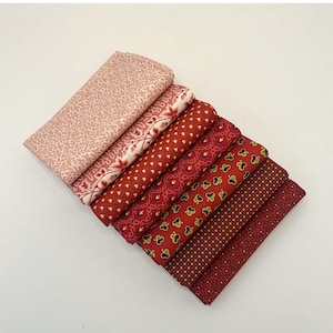 May include: A collection of folded fabric pieces in various red and pink patterns. The fabrics include floral, heart, and geometric designs. The fabrics are arranged in a diagonal row, showcasing the different patterns and colors.