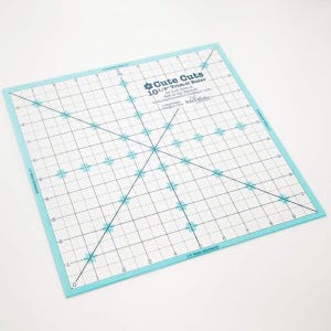 May include: A square quilting ruler with a light blue border and a grid pattern. The ruler is labeled "Cute Cuts 10 1/8" Trim-it Ruler" and features diagonal lines and measurement markings in inches. Ideal for fabric cutting and quilting.