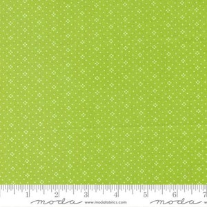 May include: Bright green fabric with a repeating pattern of small white floral shapes. The fabric is measured with a ruler in inches, showing a scale for size reference. The fabric is likely used for quilting or other sewing projects.
