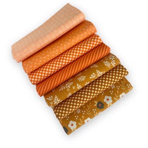 May include: A stack of seven folded fabric pieces in various shades of orange and mustard yellow. The patterns include thin grid lines, small white dots, gingham checks, diagonal stripes, and floral prints with white and dark grey flower motifs.