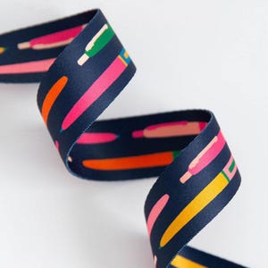 May include: A navy blue ribbon featuring a repeating pattern of colourful pencils. The pencils are in shades of pink, orange, yellow, and green. The ribbon is curled, and appears to be made of a smooth, woven material.