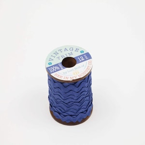 May include: A spool of blue vintage trim with a wavy edge. The spool has a wooden core and a label that reads "VINTAGE TRIM" and "BEE IN MY BONNET CO.". The trim is 12 yards long.