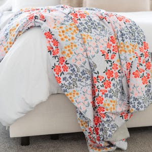 May include: A floral patterned blanket draped over a bed. The blanket features a light gray background with a colorful floral design in red, yellow, blue, and pink. The blanket is made of a soft material.