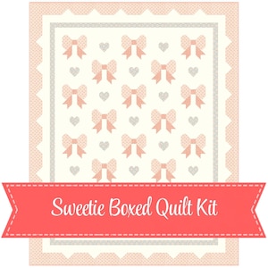 May include: A quilt kit featuring a repeating pattern of pink bows and small grey hearts on a cream background. The design is framed by a scalloped pink border with white polka dots. A coral banner across the bottom reads Sweetie Boxed Quilt Kit.