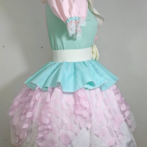 May include: A pastel-colored dress with a layered skirt. The top layer is a light blue ruffle, the middle layer is a pink lace with white polka dots, and the bottom layer is white with a scalloped edge. The dress has a white belt and a pink bow.