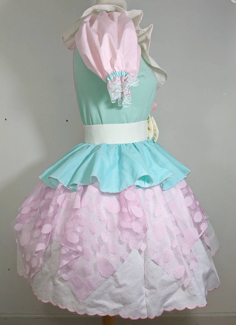 May include: A pastel-colored dress with a layered skirt. The top layer is a light blue ruffle, the middle layer is a pink lace with white polka dots, and the bottom layer is white with a scalloped edge. The dress has a white belt and a pink bow.