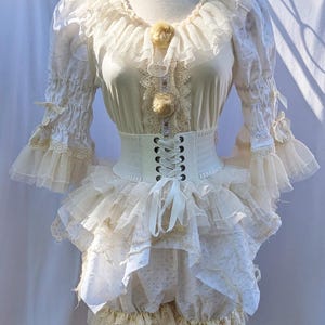 May include: Cream-colored Lolita-style dress with a corset belt. The dress features layered ruffles, lace trim, and long sleeves with ruffled cuffs. A decorative pom-pom and lace adorn the bodice. The outfit is displayed on a mannequin.