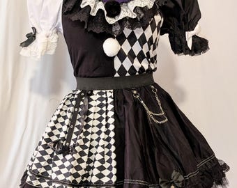 Puede incluir: Un vestido blanco y negro con patrón de rombos de arlequín, cuello de encaje con volantes y dos pompones, mangas abullonadas con lazos y una falda acampanada adornada con una cadena plateada, correas y un pequeño lazo en el dobladillo.