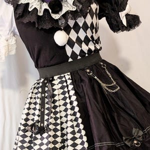 Peut inclure: Une robe noire et blanche avec un motif à losanges, un col en dentelle à volants avec des pompons, des manches courtes bouffantes avec des nœuds, et une jupe ornée d'une chaîne argentée, de détails métalliques et d'un petit nœud noir.