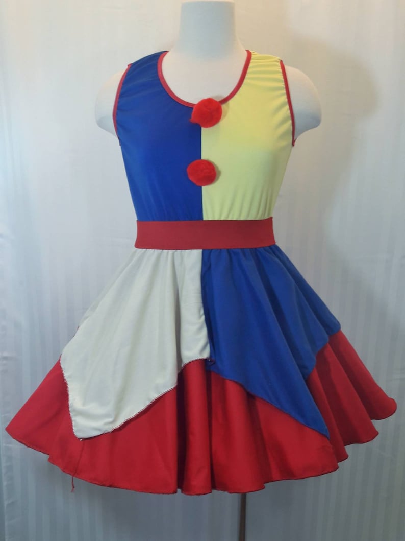 Puede incluir: Un vestido sin mangas rojo, blanco y azul con un corpi&ntilde;o amarillo y azul y una falda roja, blanca y azul. El corpi&ntilde;o tiene dos pompones rojos en la parte delantera. La falda es una falda circular con un patr&oacute;n rojo, blanco y azul.
