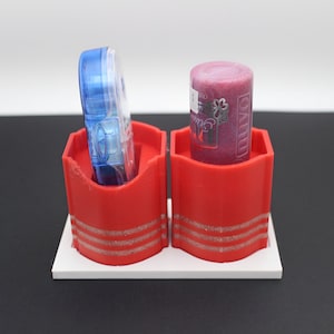 May include: A red desktop organizer with two compartments, each holding an item. One holds a blue, transparent object, and the other a cylindrical, pink item with a label. The organizer sits on a white base.