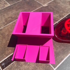 May include: A bright pink, 3D-printed organizer with two large rectangular compartments on top and three smaller compartments below. The organizer sits on a tiled surface, with a red and clear adhesive dispenser visible in the background.