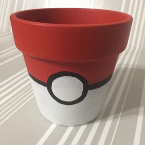 May include: A red, white, and black painted ceramic flower pot. The top of the pot is red, the bottom is white, and a black circle with a white center is painted on the front. The pot is designed to resemble a Pokeball.