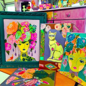 May include: A collection of colorful, vibrant paintings featuring portraits and floral designs. The artwork includes a portrait of a woman with a floral headdress, a cat wearing a mask, and other abstract floral compositions. The paintings are framed and unframed.