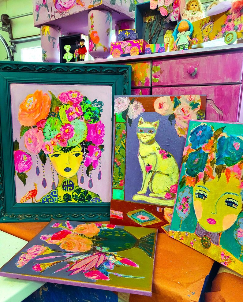 May include: A collection of colorful, vibrant paintings featuring portraits and floral designs. The artwork includes a portrait of a woman with a floral headdress, a cat wearing a mask, and other abstract floral compositions. The paintings are framed and unframed.