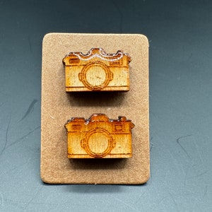May include: Two wooden camera earrings, each with a detailed design of a camera lens and body. The earrings are displayed on a brown card.