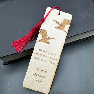 May include: Wooden bookmark with a red tassel, featuring a quote about words being as powerful as a blade. The quote reads "Words are just as capable of eviscerating someone as a blade: "It's been. My honor." - Liam"