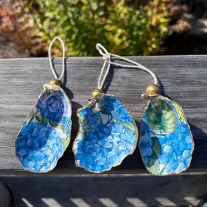 May include: Three decorative oyster shell ornaments. Each shell features a blue hydrangea floral design and a gold-painted edge. They hang from a white cord with a gold bead.