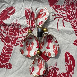 May include: Four white ceramic lobster-shaped bottle stoppers with gold accents. The stoppers are arranged in a cluster on a white fabric background with a red lobster print.