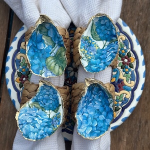 Puede incluir: Cuatro anillos de servilleta azules y dorados con un diseño floral. Los anillos están hechos de mimbre tejido y tienen un diseño de concha de ostra pintada de oro. Los anillos están sobre una servilleta blanca y un plato con estampado azul y blanco.