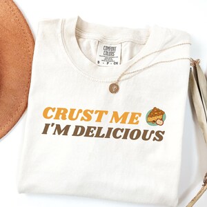 May include: A beige t-shirt with the text "CRUST ME I'M DELICIOUS" in brown and orange letters. A small illustration of a pie slice is next to the text.