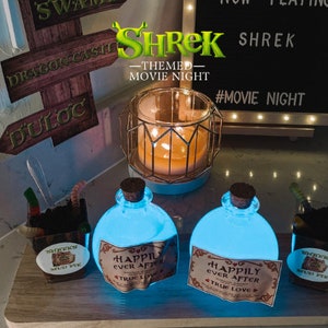 May include: A Shrek-themed movie night setup with two blue glass bottles with labels that say "Happily Ever After" and "True Love". The bottles are lit from within, creating a glowing effect. There are also two small containers of "Shrek's Mud Pie" and a candle in a gold cage.