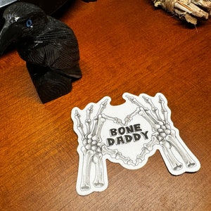 May include: White sticker with two skeleton hands forming a heart shape. The text "BONE DADDY" is inside the heart.