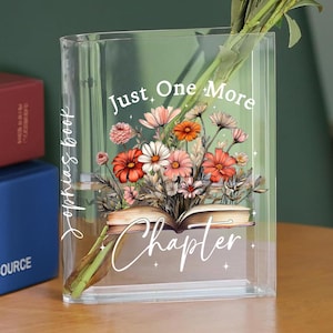 May include: A clear glass vase shaped like a book with the text "Just One More Chapter" and a floral design. The vase is filled with water and has a bouquet of flowers in it.