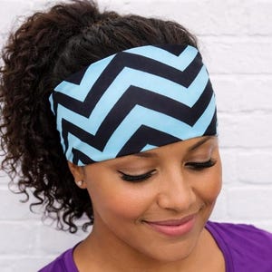 May include: A wide headband with a black and light blue chevron pattern. The headband is worn on the head and is made of a soft, stretchy material. The background is a white brick wall.
