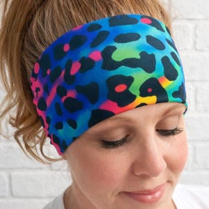 Op de afbeelding: Een kleurrijke hoofdband met een levendig luipaardprintpatroon. Het ontwerp heeft een mix van blauwe, groene, roze en gele tinten, met zwarte vlekken. Dit accessoire is ontworpen om om het hoofd te worden gedragen.