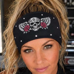May include: A black fabric headband featuring a central graphic of a white skull with detailed wings and two pink roses. The headband is embellished with small, clear, reflective rhinestones scattered across the surface of the black material.