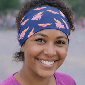 May include: A navy blue headband featuring a repeating pattern of pink pigs with yellow wings. The headband is worn by a person with dark curly hair and a purple shirt. The image is a close-up shot.