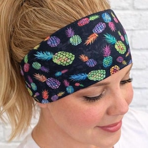 May include: Black headband with a colorful pineapple pattern. The pineapples are in shades of purple, green, yellow, and orange. The headband is worn across the forehead.