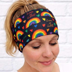 Könnte beinhalten: Schwarzes Stirnband mit Regenbogenbögen, Herzen und dem Schriftzug "Love is Love" in einem farbenfrohen Design. Das Stirnband wird von einer Person auf dem Kopf getragen, mit ihren blonden Haaren zu einem Pferdeschwanz gebunden.
