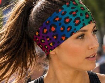 Puede incluir: Una diadema ancha de tela con un fondo de degradado arcoíris vibrante y un patrón repetitivo de estampado de leopardo negro. Los colores transicionan suavemente desde el verde en la parte superior hasta tonos azules, morados y rojos.