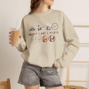 Op de afbeelding: Een beige sweater met ronde hals en een grafische print met de tekst TWENTY | ONE | PILOTS. Het ontwerp bevat diverse cirkelvormige logo's, symbolen en gestileerde afbeeldingen in zwarte, rode en blauwe tinten op de borst van het kledingstuk.