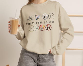 Puede incluir: Una sudadera de cuello redondo en color beige con un estampado gráfico que incluye el texto TWENTY | ONE | PILOTS. El diseño presenta varios logotipos circulares, símbolos e imágenes estilizadas en tonos negros, rojos y azules en el pecho.