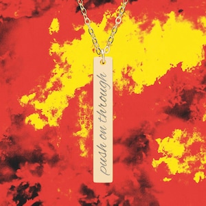 Puede incluir: Un collar dorado con un colgante rectangular con la inscripción "push on through" en cursiva. El fondo es una mezcla de colores rojo, amarillo y negro.